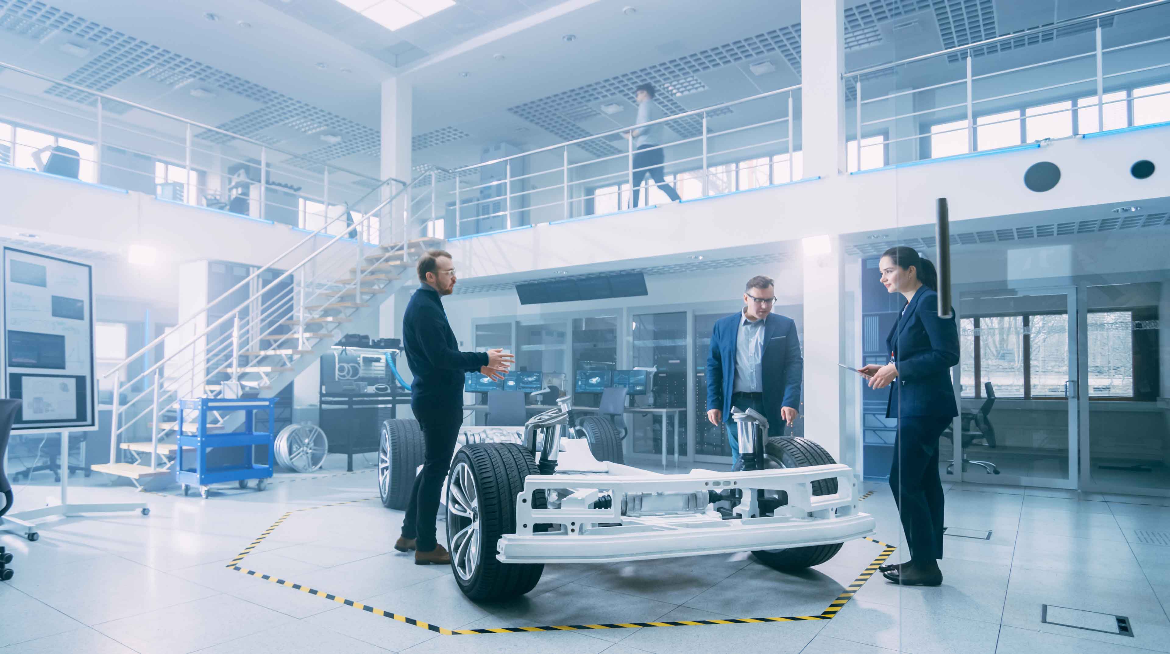 Automotive Design Engineers Talking while Working on Electric Car Chassis Prototype. In Innovation Laboratory Facility Concept Vehicle Frame Includes Wheels, Suspension, Engine and Battery.